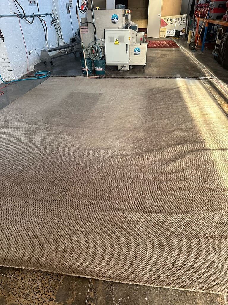 Jute rug laid out in a cleaning facility with industrial equipment and Oriental Rug Cleaning banner.