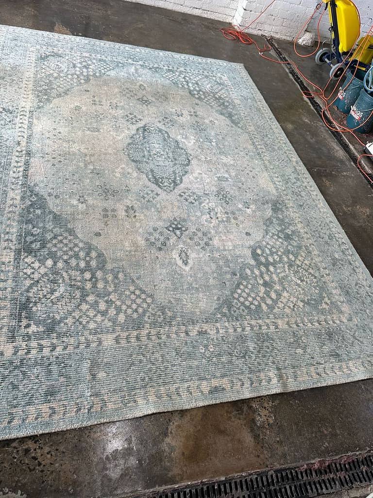 Patterned Rug in Workshop Environment Image of a large blue-gray patterned rug on a concrete floor with cleaning equipment nearby.
