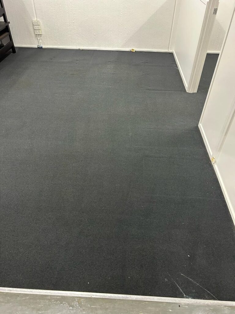 Clean Office Carpet in Factory Environment Image of a clean dark-colored office carpet in a factory setting with white walls.